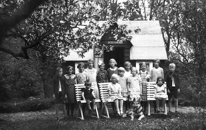Martin Svensson var ofta hos kyrkoherde Joel Nordström för att fotografera konfirmander, först i Sjörup och sedan i Gessie. Samtidigt fotograferade han prästfamiljen. Av packlådorna vid flytten till Gessie byggdes denna lekstuga, ”Gessiehus”. Martin har här fotograferat Karin Nordström och hennes kamrater när Karin fyllde 10 år 1933 (flickan med rosetten i håret). Karins kamrater är inte identifierade. Vet du vilka de är? 